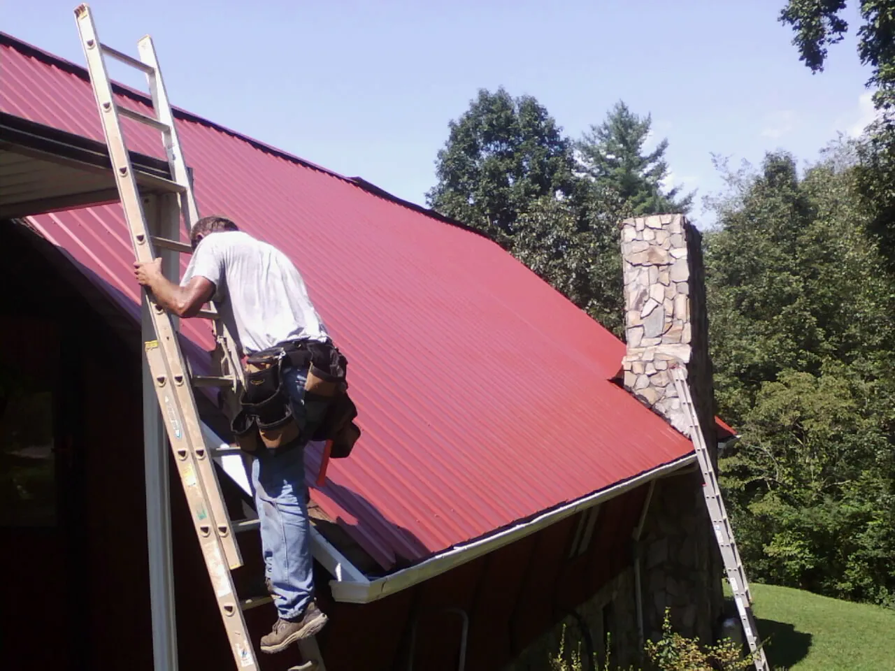 Roofer on a ladder beside a red metal roof and stone chimney — Soffit & Fascia Repair by Roofing Brothers in Fayville